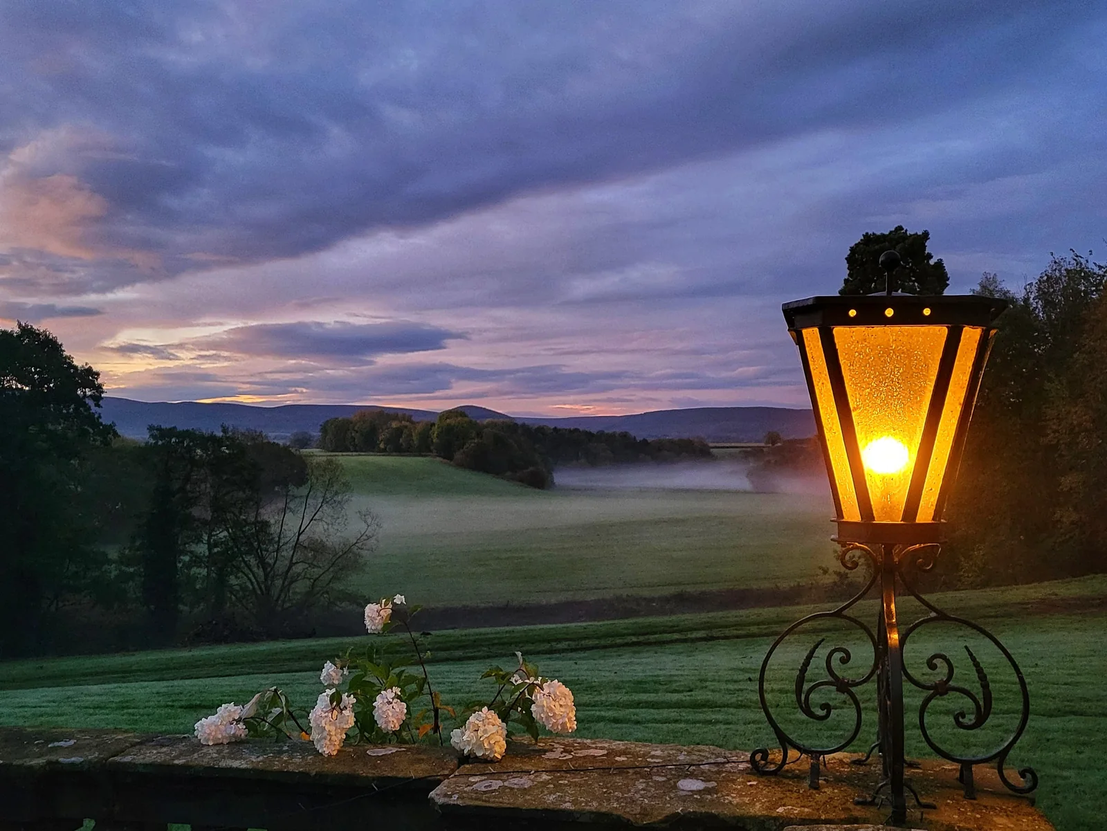 A glowing estate lamp beside flowers, with misty grounds beyond.